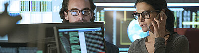 Zwei IT-Fachkräfte analysieren Daten am Monitor, eine Frau telefoniert, digitale Anzeigen im Hintergrund.