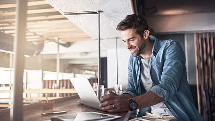 Young brunette man with a three-day beard in a blue shirt and gray shirt smiles happily at his laptop while sitting at a counter in a modern bistro and working remotely from there