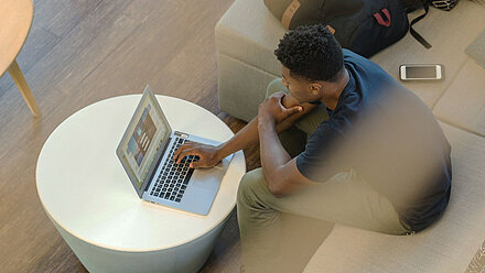 Young man sits on a sofa and works on a laptop; a smartphone lies nearby.