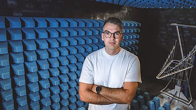 Man with glasses and folded arms stands among blue acoustic absorbers in a measurement chamber.