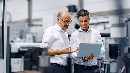 Two men in white shirts look at a laptop together in a modern industrial setting.