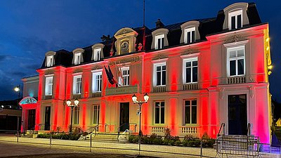 Hôtel de ville historique de nuit, illuminé en rouge et blanc, avec des drapeaux français et des lampadaires au premier plan