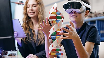 Boy wearing a VR headset reaches toward a DNA model while a woman with a tablet sits beside him.