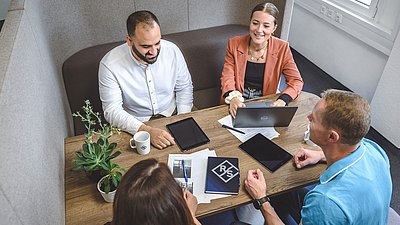 Four people discuss work at a table with a laptop, tablets and documents in a bright office.