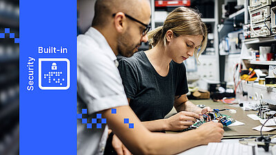 Two people work on electronic components in a workshop, with R&S®LANCOM Built-in Security shown on the left.