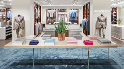 Photography of the sales area of the noble fashion store Lodenfrey: product table with elegantly arranged men's shirts in different colors and a plant in the foreground and well-stocked shelves and display walls including two men's fashion mannequins equipped with beige men's suits in the background