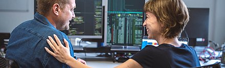Work situation with view from behind over the shoulders of two employees: Young woman in T-shirt with short, caramel-colored hair pats her blond colleague in shirt appreciatively on the shoulder; in the background, several PC screens with programming language on them can be seen on a desk