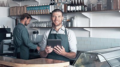 Happy barista in apron stands behind coffee and wine bar with colleagues, holding tablet in hand