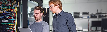 Two men stand in front of a server cabinet and discuss