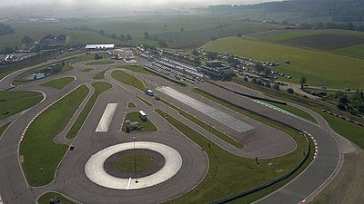 Aerial view of a vehicle test track with winding roads and a roundabout, surrounded by green fields.