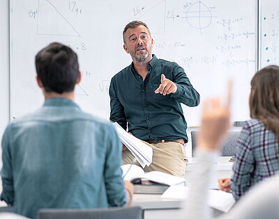 Middle-aged teacher stands in front of whiteboard with mathematical formulas and calls on student who is about to report in