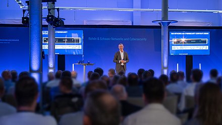 Managing Director Constantin von Reden gives a presentation on a blue-lit stage in front of partners from Rohde & Schwarz Networks and Cybersecurity