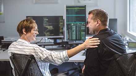 Smiling woman rests a hand on a man's shoulder at a computer workstation; monitors display programming code.