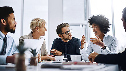 Businesspeople discuss in a bright office as a smiling woman speaks and gestures at the table.