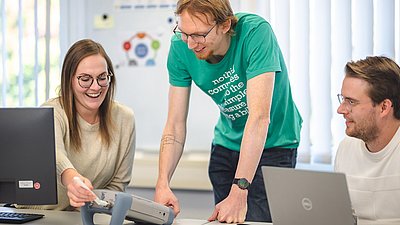 Three colleagues smile while working together on a technical device beside two laptops in a bright office.