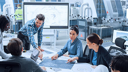 Team reviews technical plans in a modern engineering workspace with machines and monitors.