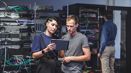Two IT specialists review a tablet beside server racks while a technician works on the network in the background.