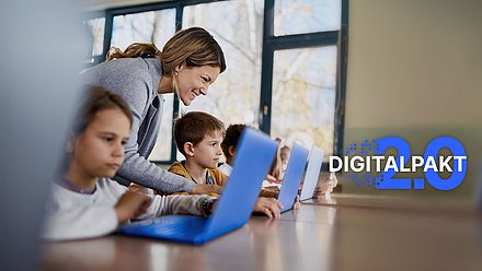Lehrerin hilft lächelnd Kindern beim Arbeiten an Laptops im Klassenzimmer, „Digitalpakt 2.0“ im Bild