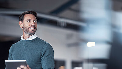 Brunette middle-aged man with a beard and a combination of a blue sweater over a white shirt on the left of the picture stands smiling confidently in modern office space, holding a tablet in his hand and looking inspired and thoughtful up to the right