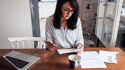 Young brunette woman with glasses sits at home at dining table with kitchen in background and tries to make a bureaucratic application with laptop, printouts, and cell phone