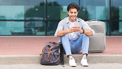 Traveling man sits with smartphone and suitcase on sidewalk in front of airport concourse