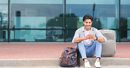 Traveling man sits with smartphone and suitcase on sidewalk in front of airport concourse