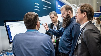Vier Männer diskutieren auf einer Fachmesse gemeinsam die Darstellung auf einem großen Bildschirm.